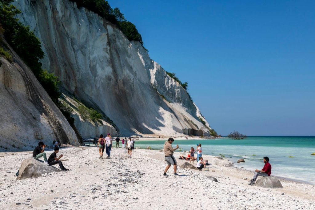 하얀 절벽이 있는 해변, 경치를 즐기며 흩어져 있는 방문객, 맑고 푸른 하늘 아래 잔잔한 청록색 바다.