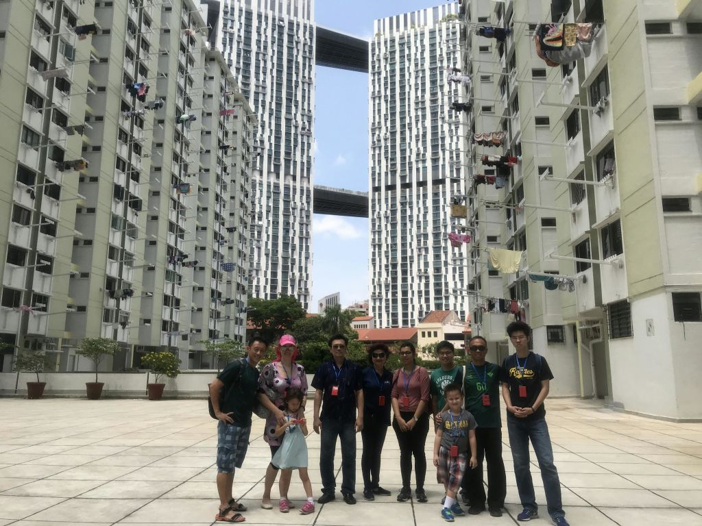 한 무리의 사람들이 하늘 다리가 있는 두 개의 고층 아파트 건물 사이에 야외에 서 있으며, 발코니에는 빨래방이 걸려 있습니다.