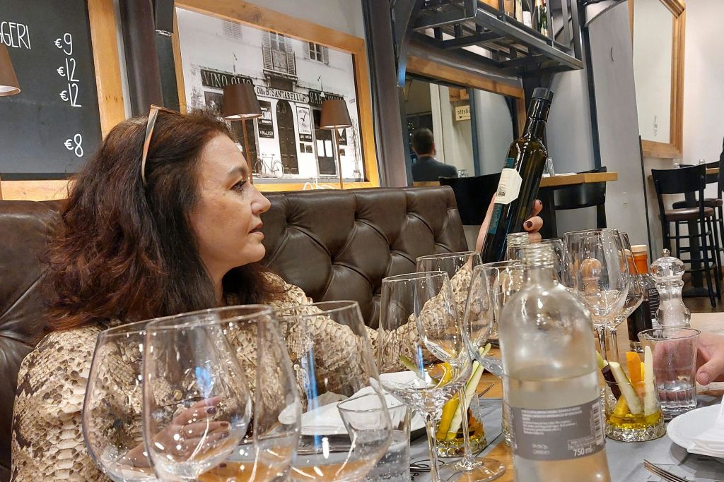 빈 와인잔과 도금된 식기류로 둘러싸인 레스토랑 테이블에 앉아 있는 여성으로, 메뉴 가격을 배경으로 볼 수 있습니다.