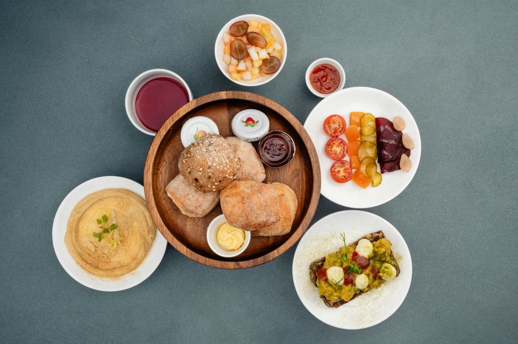 빵 롤, 후무스, 구운 야채, 과일 샐러드, 소스, 열린 얼굴의 샌드위치를 포함한 다양한 요리가 테이블에 있습니다.