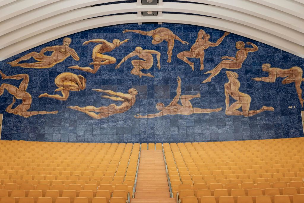 노란색 좌석이 줄지어 있고 파란색 배경에 사람 형상이 떠 있는 커다란 벽화가 있는 강의실.