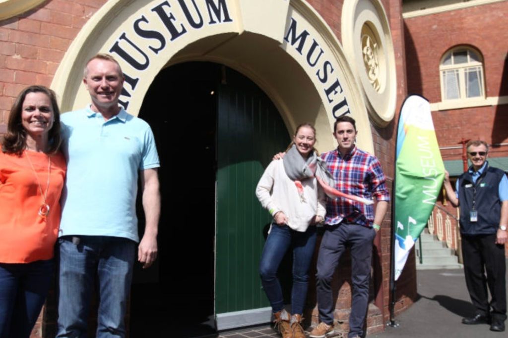 화창한 날 근처에 직원과 깃발이 있는 박물관 입구 앞에서 두 사람이 미소를 지으며 서 있습니다.