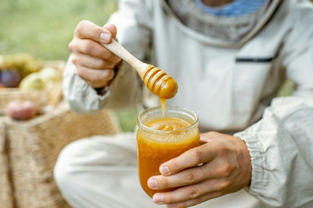 양봉복을 입은 사람이 벌꿀 항아리를 들고 벌꿀 디퍼를 야외에 담그고 있습니다.