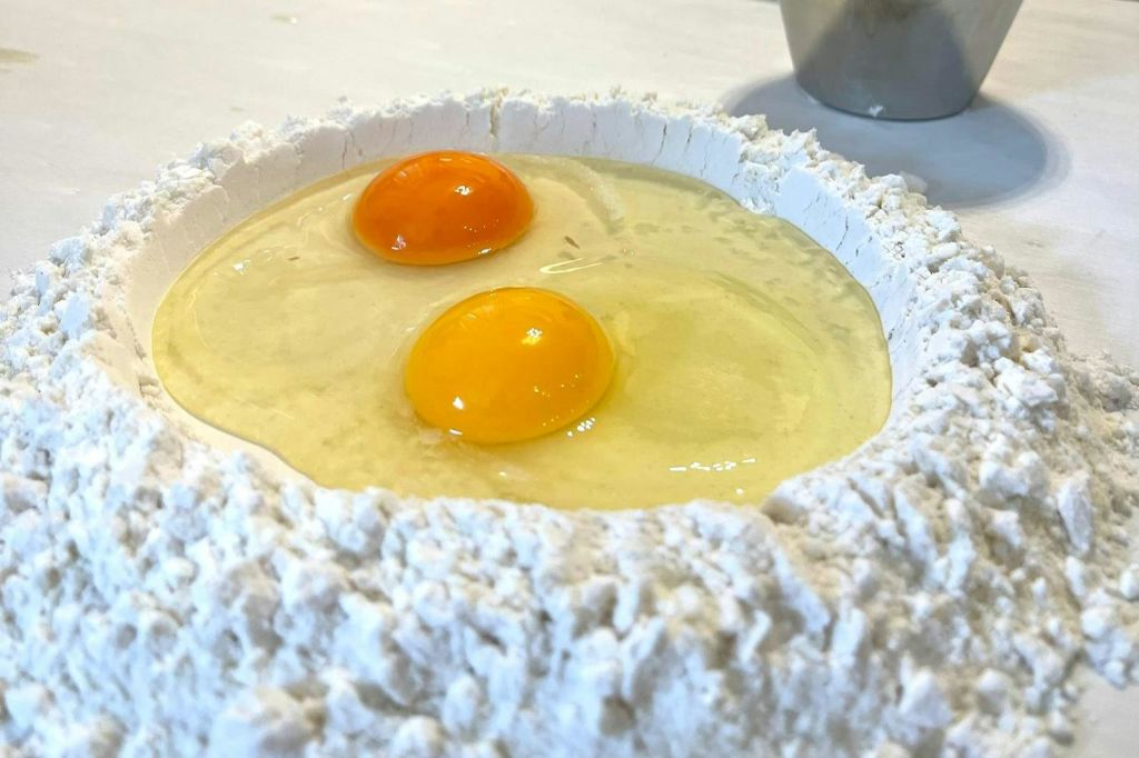 두 개의 날계란이 조리대 위에 있는 밀가루 우물에 금이 갔고, 금속 컵이 배경에 부분적으로 보였다.