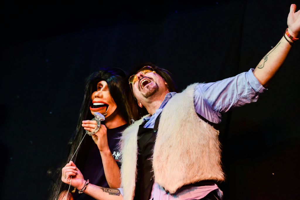 모피 조끼와 선글라스를 착용한 사람이 무대에서 짙은 머리의 복화술사 더미를 들고 공연을 합니다.