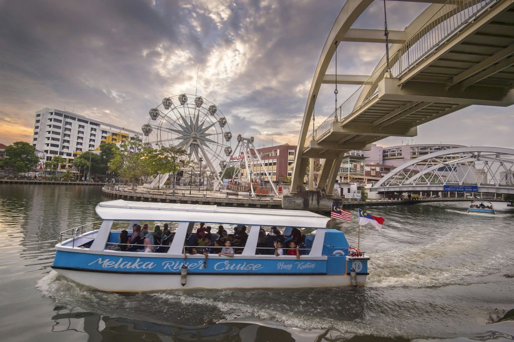 다리 아래에는 "멜라카 강 크루즈" 라고 쓰여진 보트가 있습니다. 관람차와 건물이 배경에 있습니다.