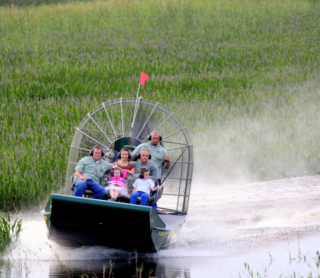 어린 소녀와 소년을 포함한 6명이 에어보트를 타고 잔디가 우거진 습지를 통과합니다. 프로펠러와 녹지가 보입니다.