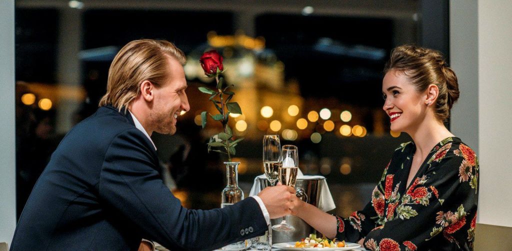 꽃무늬 드레스를 입은 한 여성이 보케 조명이 있는 어두운 분위기에서 저녁 식사 테이블 위로 다른 사람과 함께 미소를 짓고 안경을 핥고 있습니다.