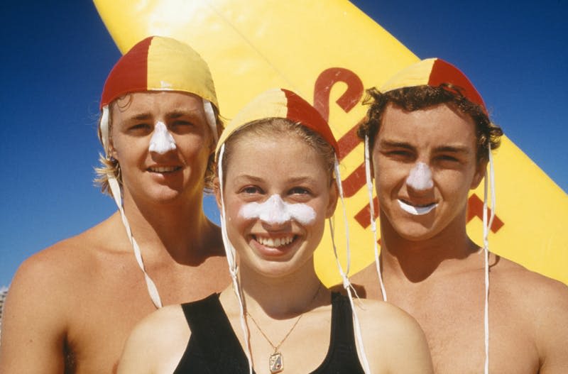 빨간색과 노란색 수영모를 쓰고 코에 아연을 바른 세 사람이 노란색 서핑보드 앞에 서 있습니다.