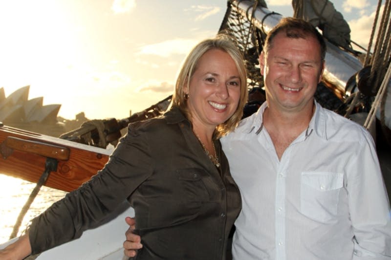 해가 질 무렵 밧줄과 돛이 달린 범선 앞에서 미소를 짓는 남녀.