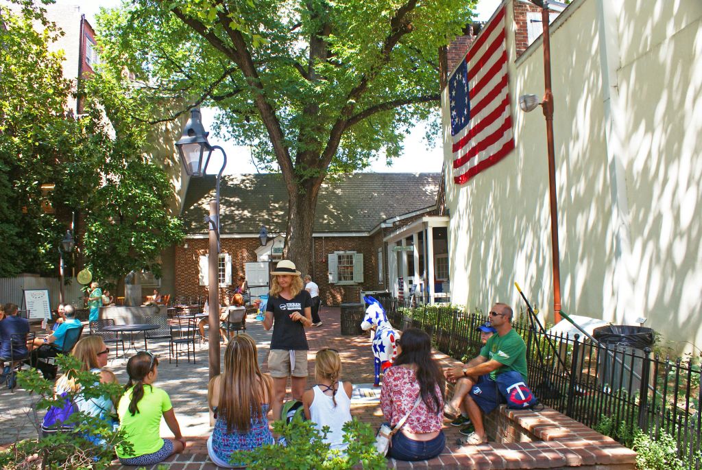 벽돌 건물과 미국 국기 옆 그늘진 마당에 한 무리의 사람들이 앉아 서 있습니다.