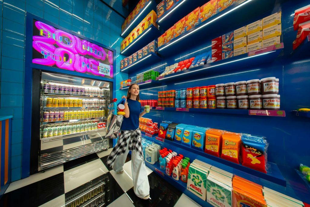 화려한 색상의 선반과 네온 사인이 있는 밝은 조명의 매장에서 한 사람이 쇼핑백과 음료를 들고 걸어가고 있습니다.