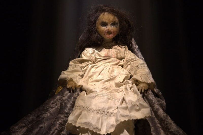 검은 머리카락과 흰색 드레스를 입은 마모된 인형으로, 극적인 조명이 있는 어두운 드레이프 배경에 앉아 있습니다.