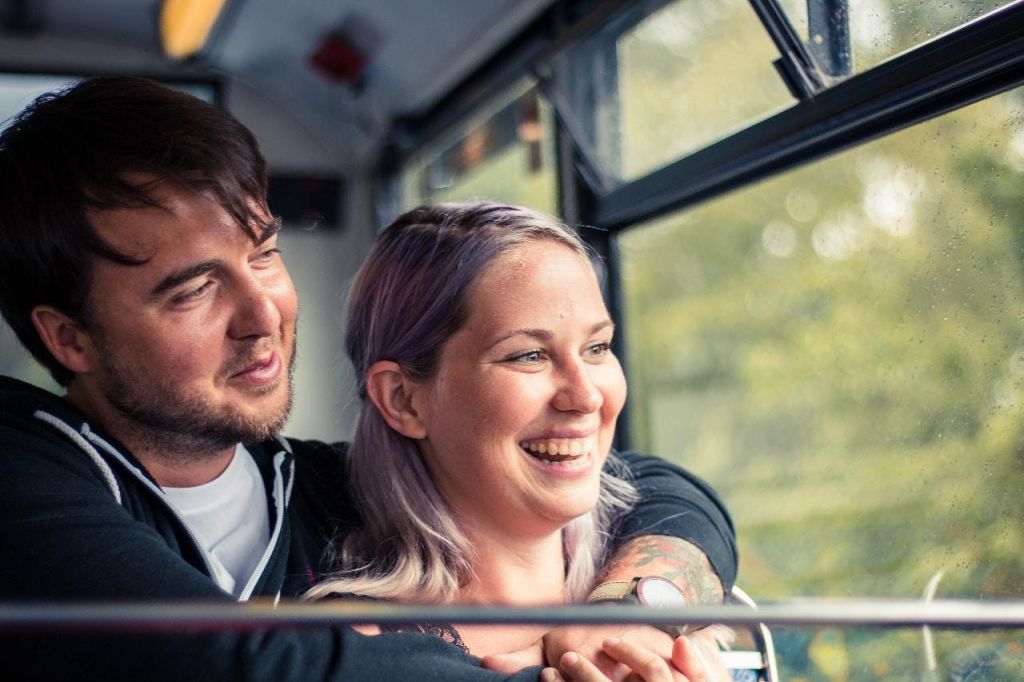버스 안에서 한 남자와 여자가 포옹하고 미소를 짓고 있으며, 창문과 조명이 희미한 내부가 배경으로 보입니다.