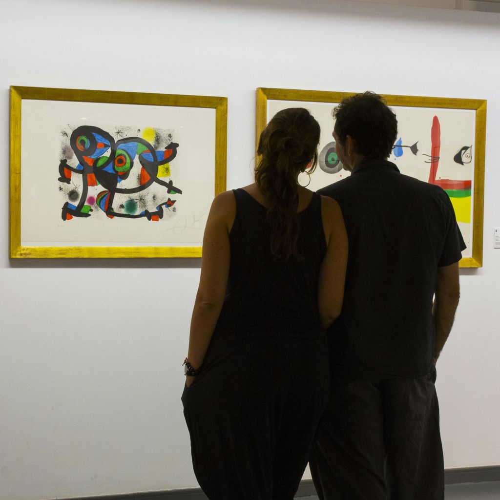 두 사람이 가까이 서서 흰색 갤러리 벽에 금색 액자로 장식된 추상 예술 작품을 감상하고 있습니다.