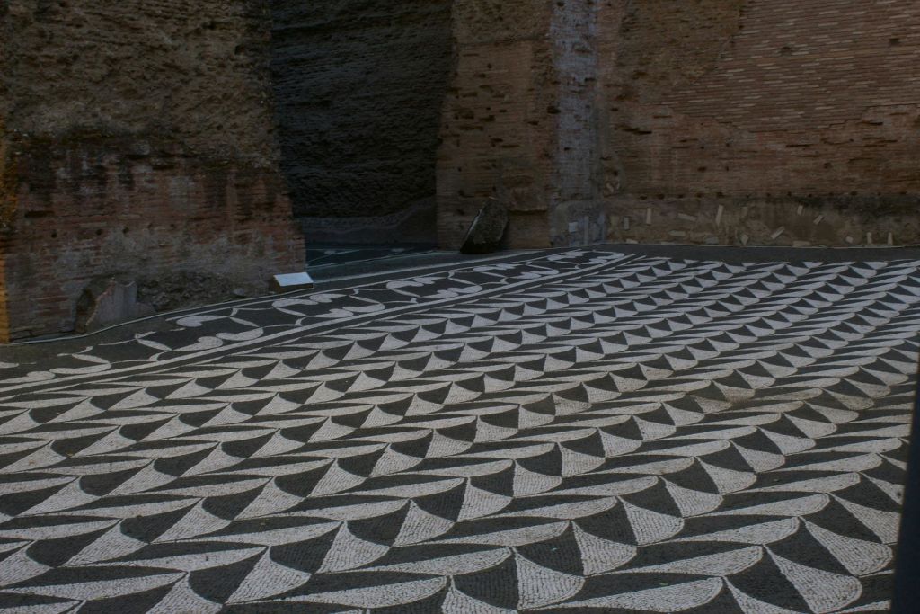 거친 갈색 벽돌 벽 옆에 흑백의 기하학적 모자이크 바닥 패턴이 있고 바닥에 작은 표지판이 있습니다.