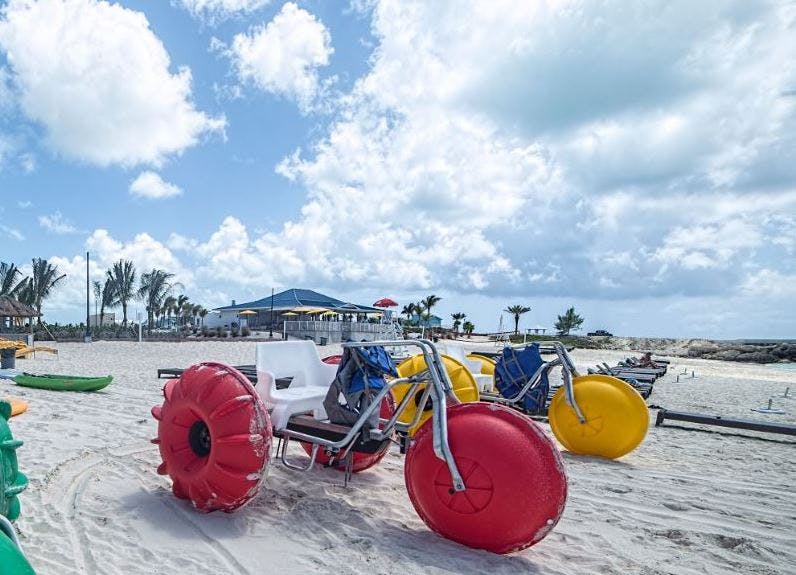 부분적으로 흐린 하늘 아래 모래사장에 커다란 빨간색과 노란색 바퀴가 달린 대형 세발자전거가 있는 해변 풍경.