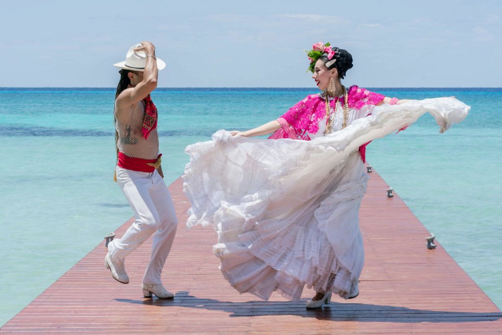 전통 복장을 한 여성이 청록색 바다 옆 나무 부두에서 활기차게 춤을 추고 있으며, 옆에는 다른 사람의 일부가 보입니다.