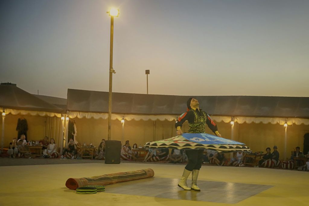 저녁 하늘 아래 야외 공연장에서 구경꾼들로 둘러싸인 전통 복장을 한 공연자가 돌고 있습니다.