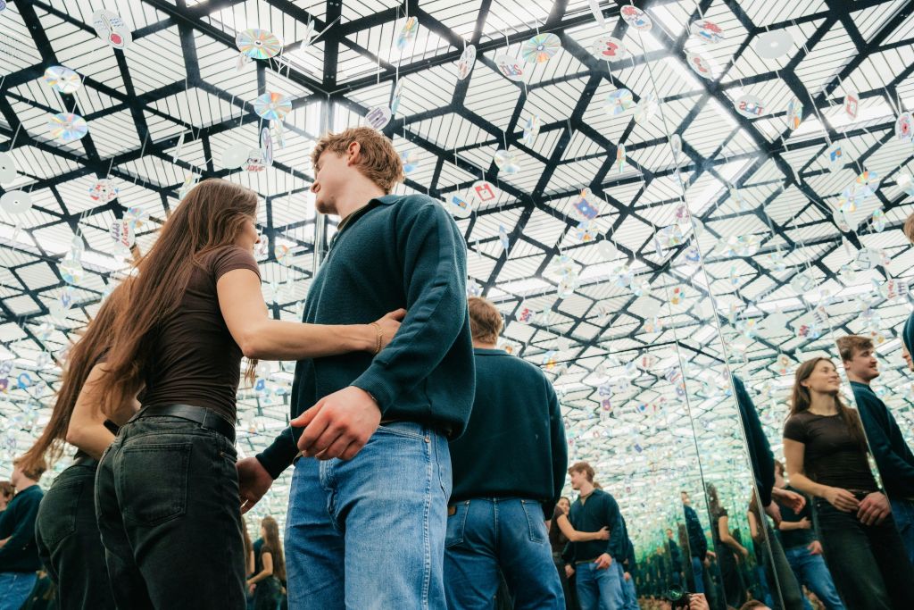 천장에 기하학적 패턴과 그림이 반영된 거울 벽이 있는 방에 서 있는 두 사람.