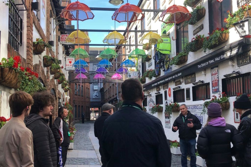 화려한 우산이 걸려 있고 장식된 벽이 있는 골목에서 한 무리의 사람들과 이야기하는 투어 가이드.