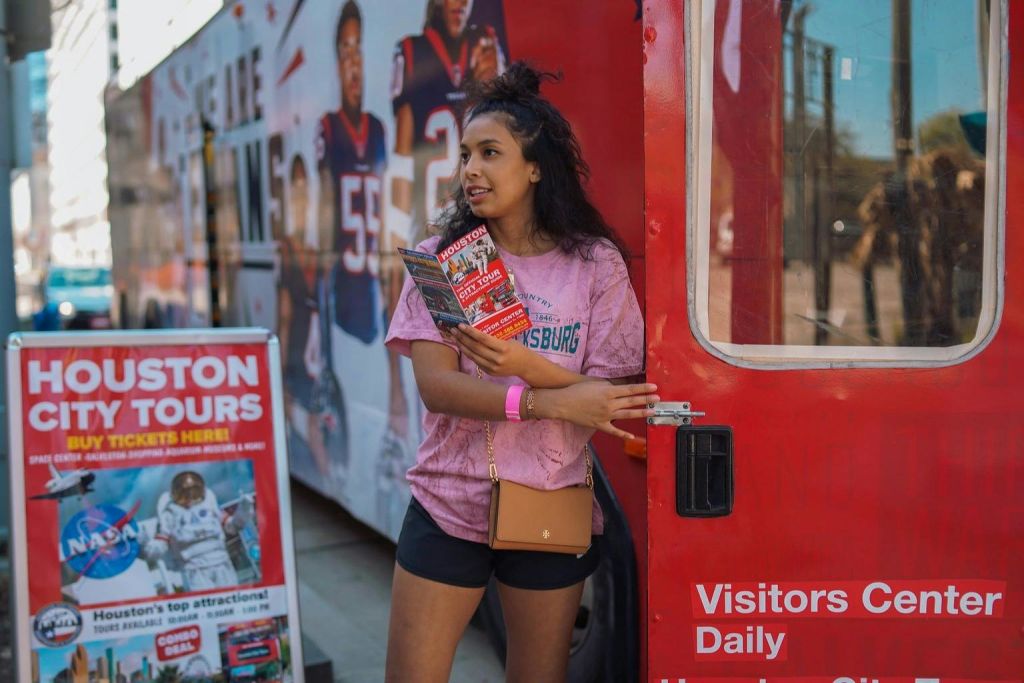 책자를 들고 있는 분홍색 셔츠를 입은 여성이 홍보 표지판과 포스터를 배경으로 빨간색 버스 근처에 서 있습니다.