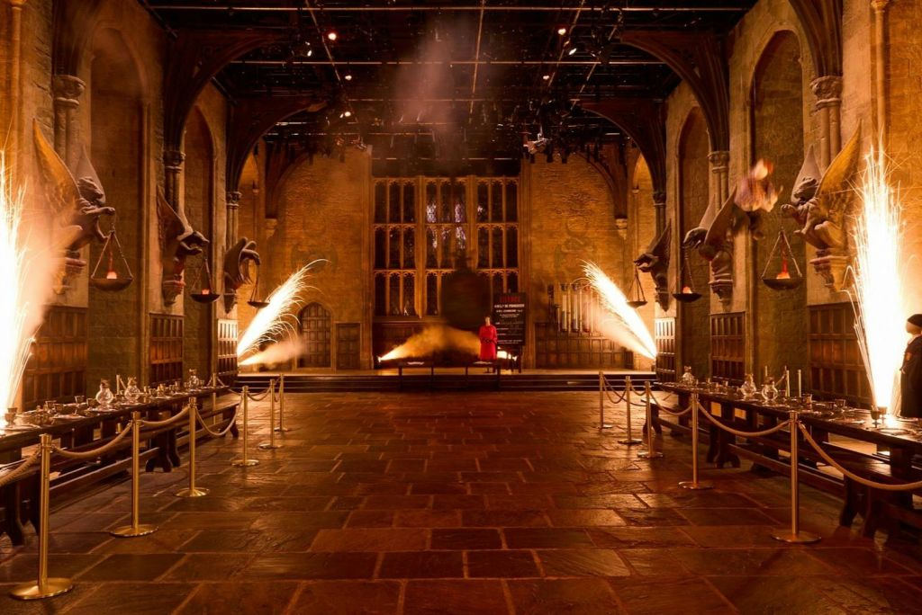 중세 건축물, 벽에 불이 켜진 횃불, 중앙 벽난로 옆에 서 있는 빨간색 인물이 있는 웅장한 홀입니다.