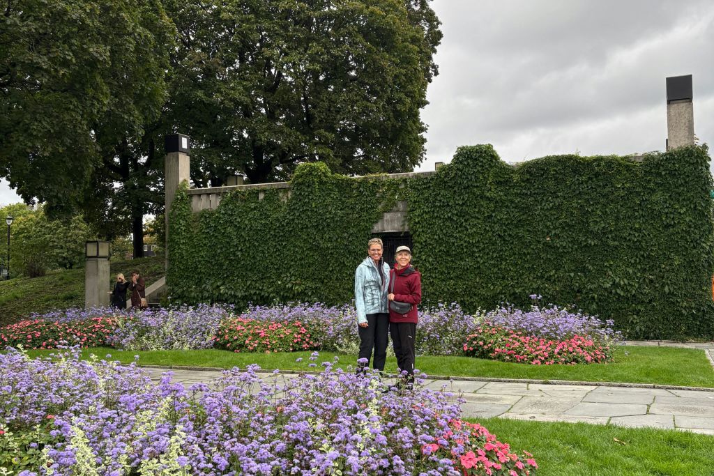 푸른 담쟁이로 뒤덮인 벽 앞에 보라색과 분홍색 꽃이 만발한 정원의 길에 서 있는 두 사람. 흐린 하늘.