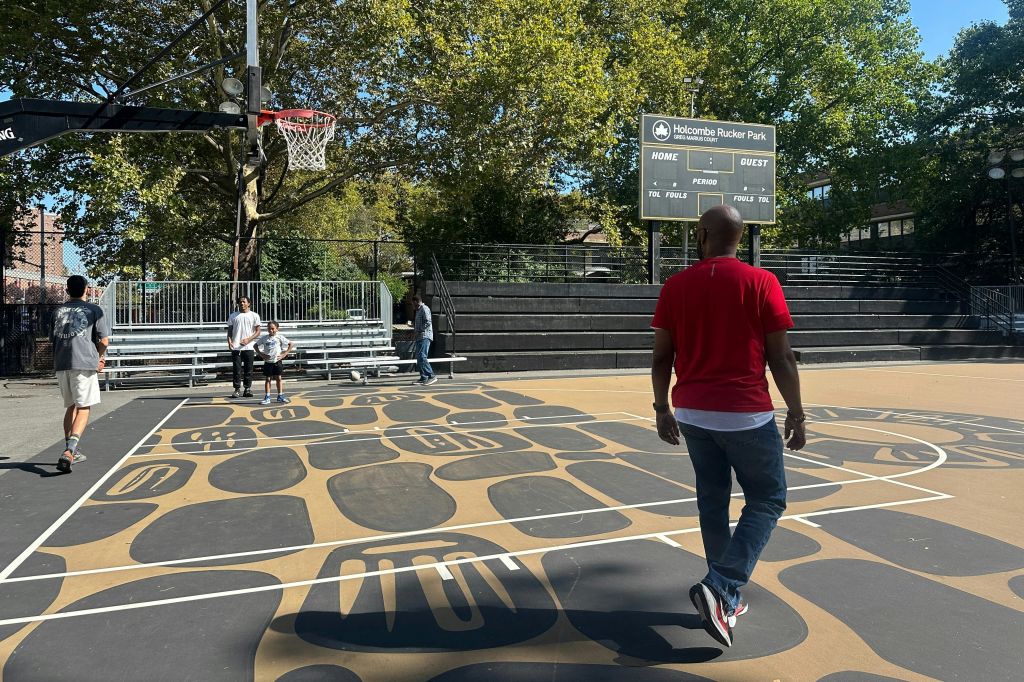 빨간 셔츠를 입은 남성이 배경에 세 사람이 보이는 점수판이 있는 패턴이 있는 야외 농구 코트를 걷고 있습니다.