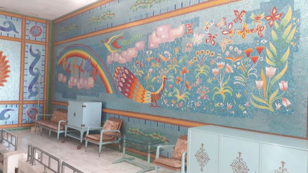 푸른 벽에 공작새, 꽃, 무지개가 그려진 다채로운 벽화 아래에는 연한 파란색 수납장과 의자가 배치되어 있습니다.