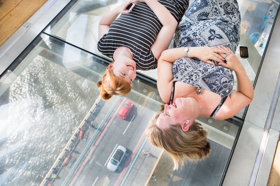 유리 바닥에 누워 서로를 바라보는 두 여성이 아래 도로와 자동차를 바라보고 있습니다.