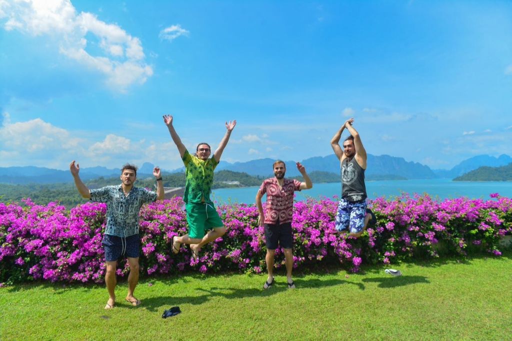 화려한 색상의 셔츠를 입은 네 명의 남성이 꽃이 피는 분홍색 꽃 앞에서 포즈를 취하고 있으며, 두 개의 점프와 경치 좋은 산과 호수를 배경으로 하고 있습니다.