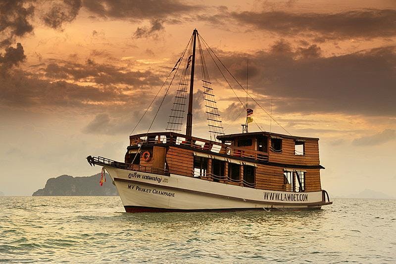 "M.V. 푸켓 샴페인" 이라는 이름의 나무 보트가 석양을 배경으로 잔잔한 물 위를 항해합니다.