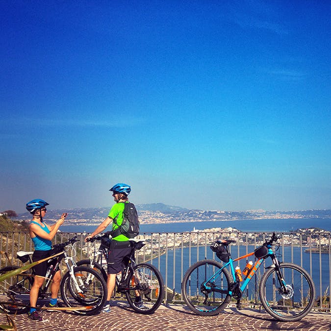 헬멧을 쓴 두 명의 자전거 운전자가 전망대에서 도시 전망을 감상하기 위해 멈춥니다. 하늘과 물을 배경으로 자전거 두 대가 나란히 서 있습니다.