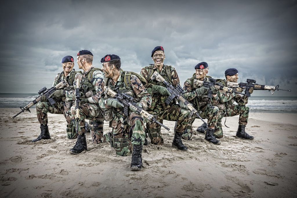 위장복과 베레모를 입은 여섯 명의 군인이 소총을 들고 흐린 하늘 아래 모래 해변에서 웅크리고 웃고 있습니다.