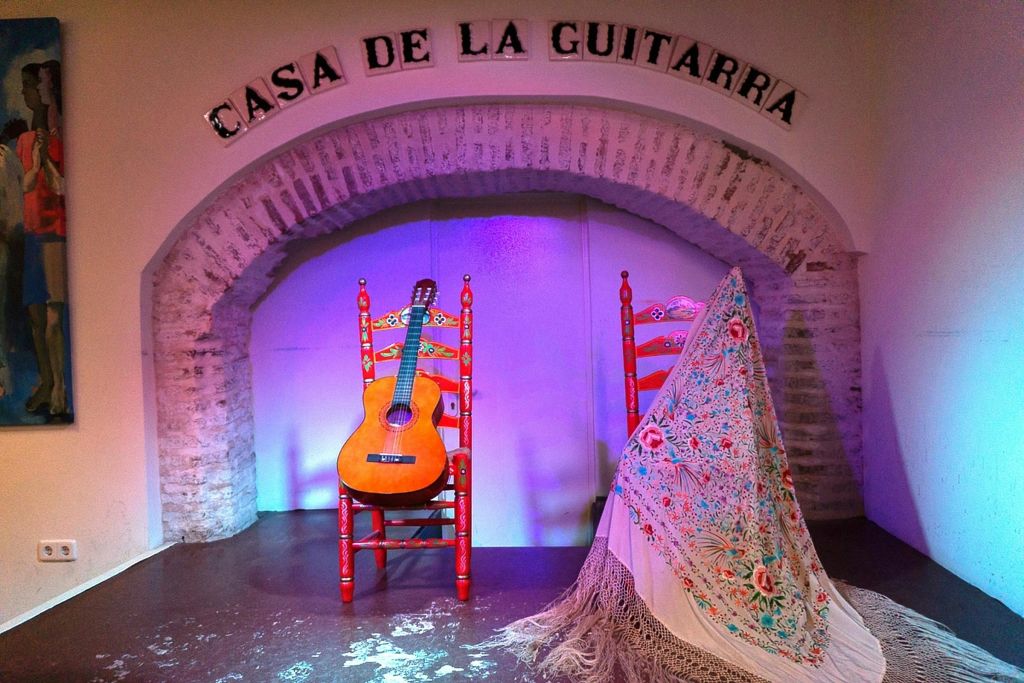 화려한 빨간색 의자에 기타가 있고 화려한 숄로 장식된 의자가 하나 더 있는 "Casa de la Guitarra" 라는 표지판이 있는 무대입니다.