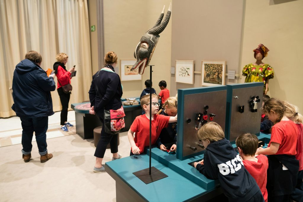 성인이 지켜보는 동안 어린이들은 박물관에서 교육 전시물과 상호 작용합니다. 부족 마스크가 스탠드에 눈에 띄게 표시됩니다.