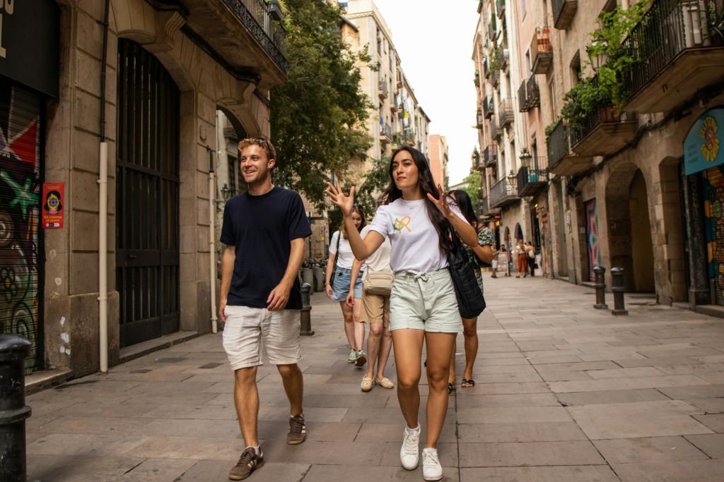 발코니와 상점이 있는 복층 건물 옆으로 가로수가 늘어선 좁은 거리를 걷고 이야기하는 사람들.