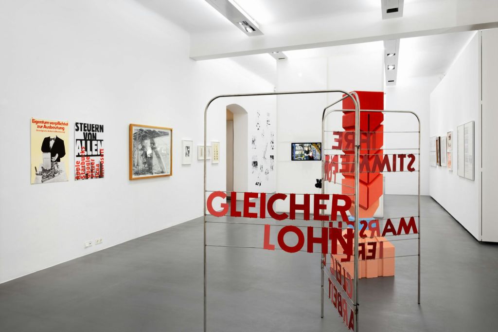 흰색 벽에 다양한 액자의 예술품과 빨간색 텍스트 "GLEICHER LOHN" 이 있는 금속 구조물이 있는 아트 갤러리입니다.