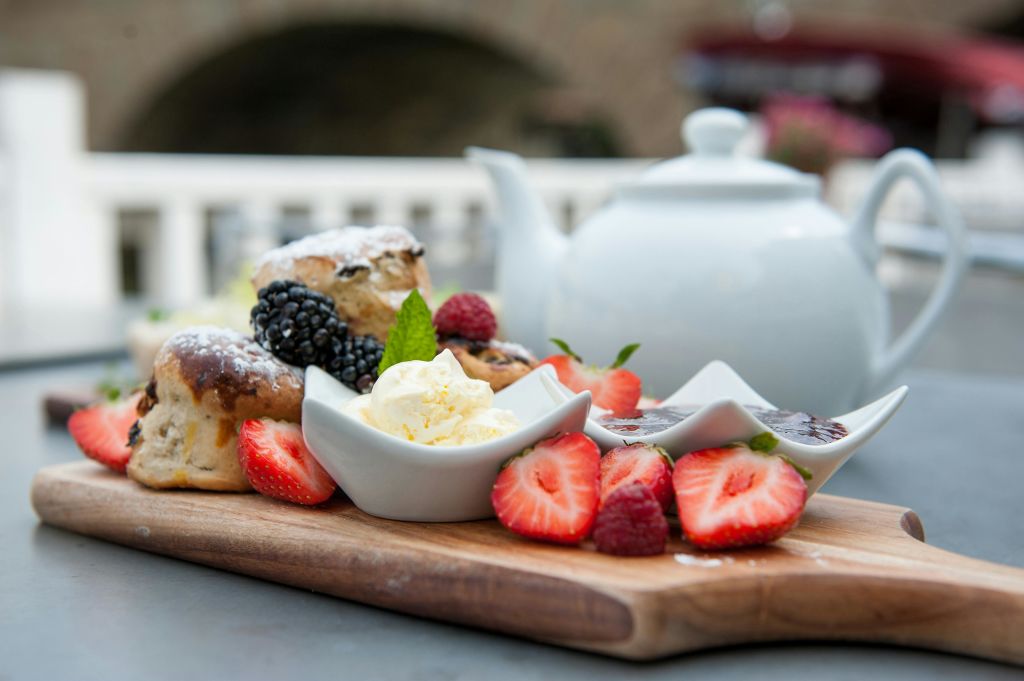 스콘, 신선한 딸기, 블랙베리, 라즈베리, 작은 크림 접시가 있는 나무판입니다. 배경에는 주전자가 있습니다.