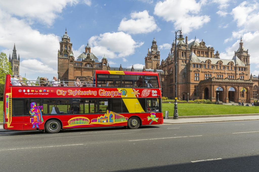 "City Sightseeing Glasgow" 가 있는 빨간색 2층 관광 버스가 부분적으로 흐린 하늘 아래 역사적인 건물을 지나갑니다.