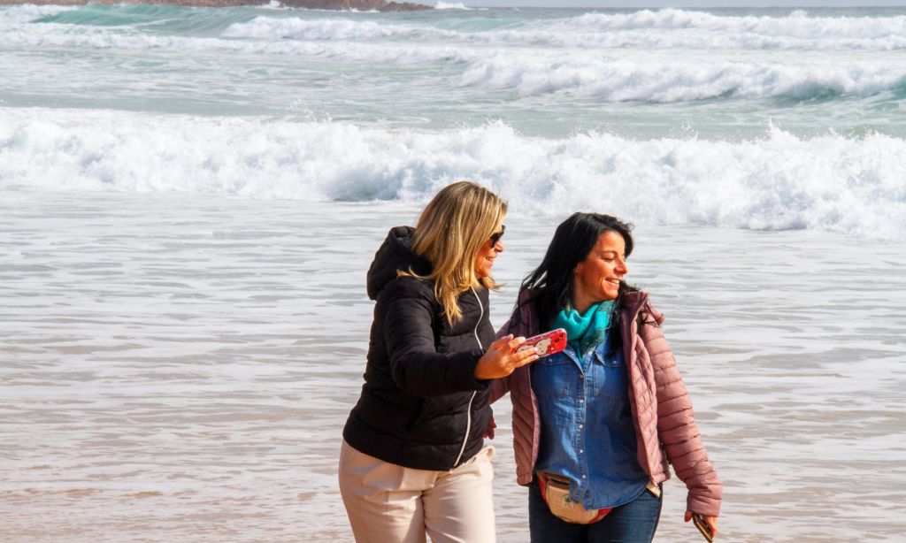 파도를 배경으로 해변을 거닐며 미소 짓는 두 여성. 한 여성이 셀카를 찍고 있습니다.