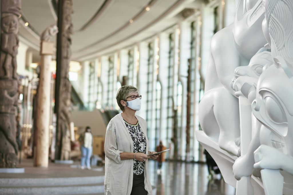 높은 천장과 커다란 창문이 있는 넓고 현대적인 갤러리에 가면을 쓴 한 여성이 커다란 조형물을 관찰하고 있습니다.