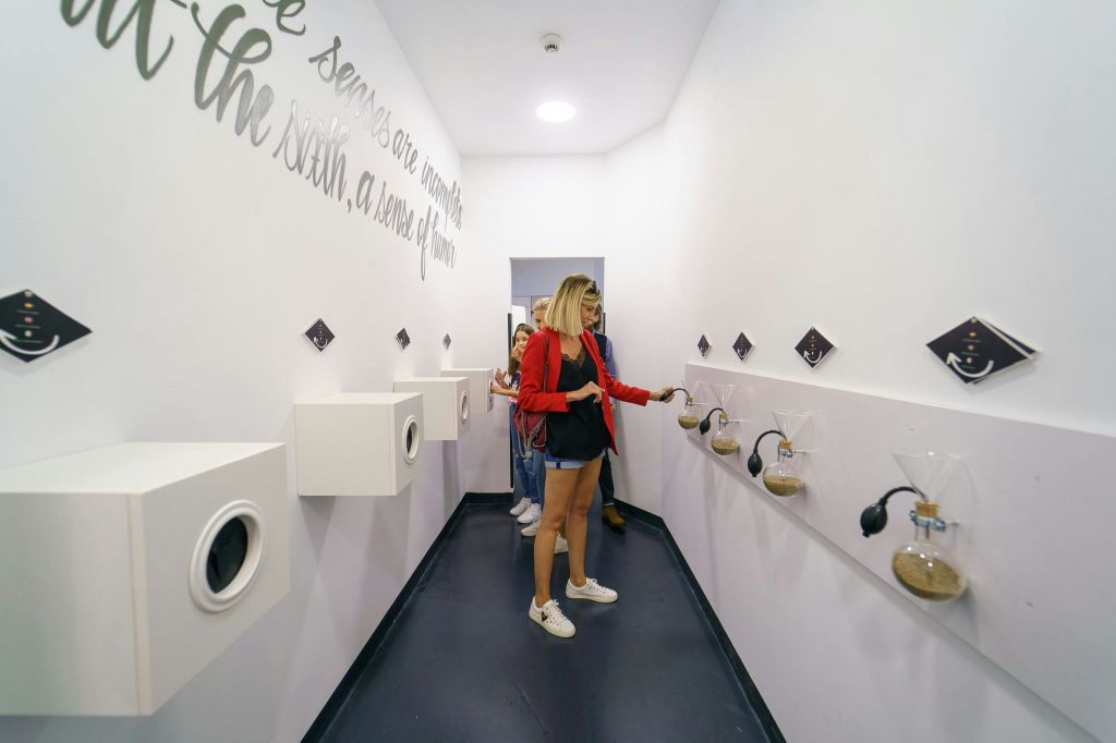 흰색 방에 있는 사람들은 벽에 설치된 다양한 냄새 스테이션과 상호 작용하며, 일부는 고무 전구를 사용하여 냄새를 냅니다.