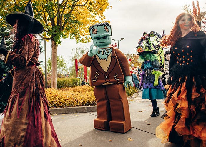 프랑켄슈타인 같은 레고 피규어를 포함한 할로윈 테마 의상을 입은 사람들이 가을 나무가 있는 공원 통로를 걷고 있습니다.