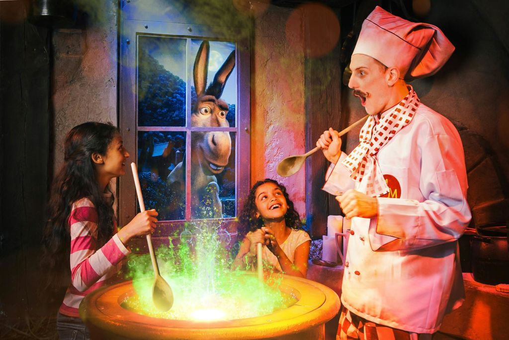 두 아이와 셰프가 녹색 액체가 든 가마솥을 휘젓고, 당나귀가 창문을 들여다보며 미소를 짓고 있습니다.