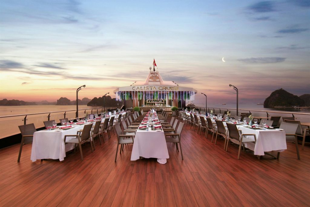 초승달이 내리쬐는 황혼 하늘 아래, 깔끔하게 정리된 테이블과 의자가 있는 유람선 데크의 야외 식사 공간.