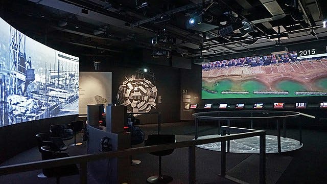 인터랙티브 디스플레이와 도시 경관과 해안선의 커다란 벽면 투영을 특징으로 하는 어두운 조명 전시실입니다.
