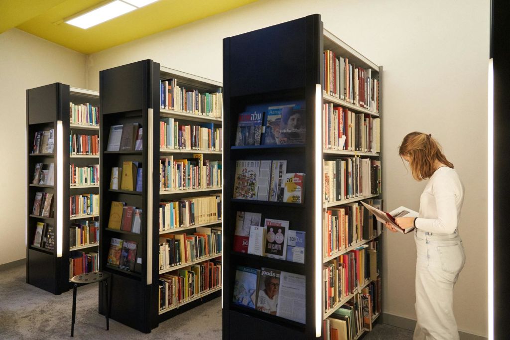 조명이 밝은 도서관에서 한 여성이 책과 잡지가 가득한 선반 옆에서 책을 읽고 있습니다.