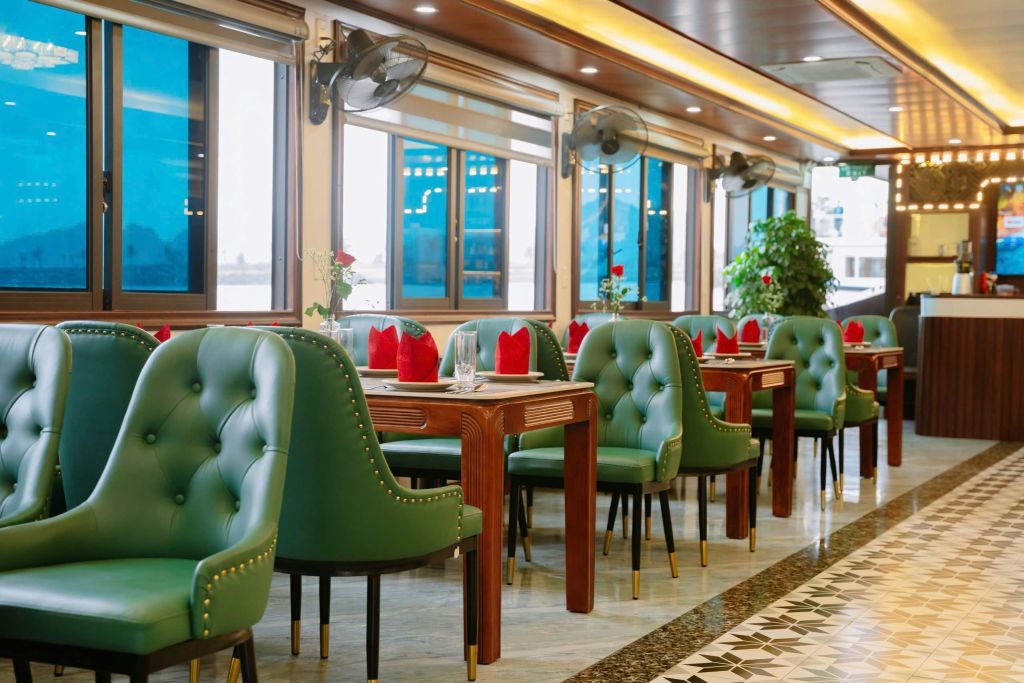 녹색 터프트 의자, 빨간 냅킨이 놓인 나무 테이블, 커다란 창문, 천장 선풍기, 화분이 있는 레스토랑입니다.